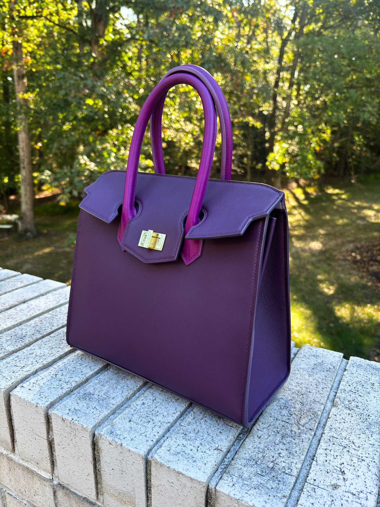 Image shows the Orion 27cm in Aubergine and Anemone. This is a trapezoid shaped handbag with a top flap closure and a gold colored locking mechanism. The handles of the bag are fixed upright and are anemone in color. Aubergine is a darker and richer purple color. Anemone is a lighter, almost magenta color purple. The edge paint is color matched to the aubergine base color of the bag.